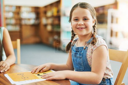 Téléchargez les photos : Je suis prêt à apprendre. Portrait d'une petite fille joyeuse faisant son travail scolaire tout en étant assise dans une salle de classe à l'intérieur pendant la journée - en image libre de droit