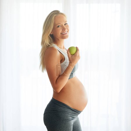 Téléchargez les photos : Certains appellent ça manger sainement, j'appelle ça aimer. Portrait d'une heureuse femme enceinte vêtue de vêtements de sport et mangeant une pomme à la maison - en image libre de droit