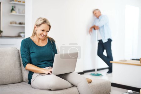 Téléchargez les photos : C'est son jour pour faire le ménage. une femme mûre utilisant un ordinateur portable à la maison pendant que son mari balaie en arrière-plan - en image libre de droit