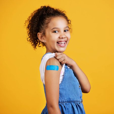 Photo for Happy, portrait and child with a plaster in a studio for a wound, sore or injury on her arm. Happiness, smile and healthy girl kid model posing with a bandaid after a vaccination by yellow background. - Royalty Free Image