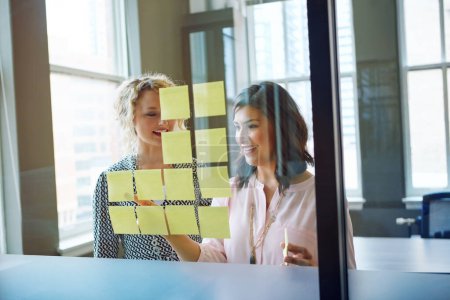 Photo for Theyre success is built on teamwork. two businesswomen brainstorming with adhesive notes on a glass wall in an office - Royalty Free Image
