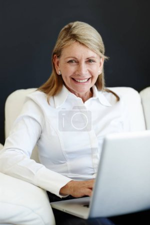 Téléchargez les photos : Prendre le temps de rester en contact. Portrait d'une femme mûre souriante assise sur un canapé à l'aide d'un ordinateur portable - en image libre de droit