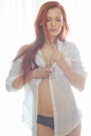 Téléchargez les photos : Portrait d'une jolie femme rousse portant une chemise déboutonnée debout dans une chambre à coucher à l'air sexy tout en se tenant debout et son déboutonnage de sa chemise. Jeune femme posant semi nue. - en image libre de droit