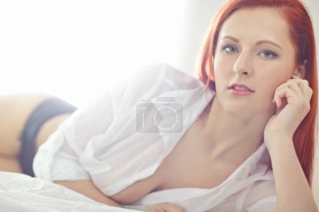 Téléchargez les photos : Portrait d'une belle jeune rousse se relaxant sur un lit à la maison. Sentiment féminin confiant flirt, sensuel et sexy tout en posant séduisante en chemise blanche déboutonnée et sous-vêtements. - en image libre de droit