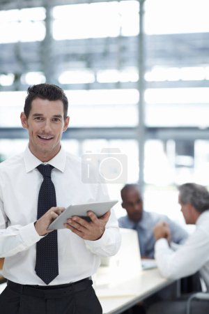 Foto de Negocios con facilidad. Un hombre de negocios guapo sosteniendo un touchpad sonriendo y mirando a la cámara con colegas trabajando en el fondo - Imagen libre de derechos