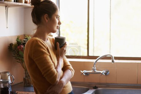 Photo for Mornings are when I miss you most. Attractive young woman in her kitchen having a warm beverage while she looks out of her window - Royalty Free Image