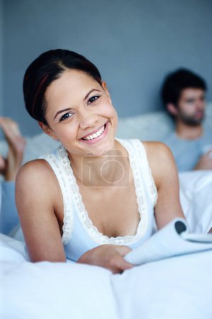 Foto de Obtener mi dosis diaria de las redes sociales. Retrato de una mujer sonriente acostada en su cama con una revista frente a ella con su novio en el fondo - Imagen libre de derechos