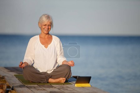Foto de Elegir un entrenamiento de yoga en línea. Una mujer madura haciendo ejercicios de yoga en un muelle con la ayuda de su tableta digital - Imagen libre de derechos