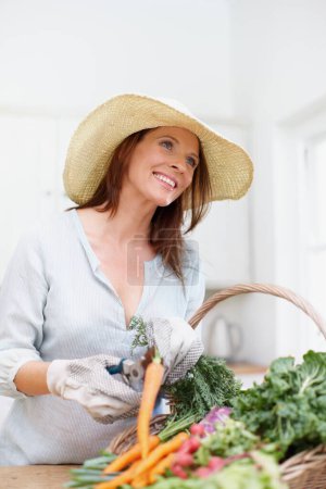 Foto de La vida sencilla. Una mujer hermosa, con un sombrero de paja, corta los tallos de verduras frescas en una cesta en el mostrador de la cocina - Imagen libre de derechos