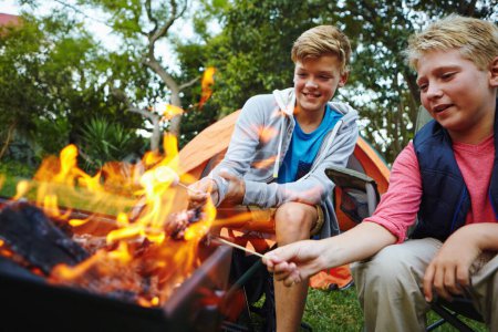 Téléchargez les photos : Ça va avoir bon goût. deux jeunes garçons assis près du feu de camp - en image libre de droit