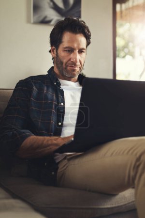 Foto de Esto es muy entretenido. un joven guapo usando su portátil mientras se relaja en un sofá en casa - Imagen libre de derechos