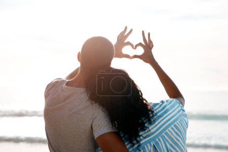 Foto de Ha sido nada menos que un día perfecto. Vista trasera de una joven pareja haciendo una forma de corazón con sus manos en la playa - Imagen libre de derechos