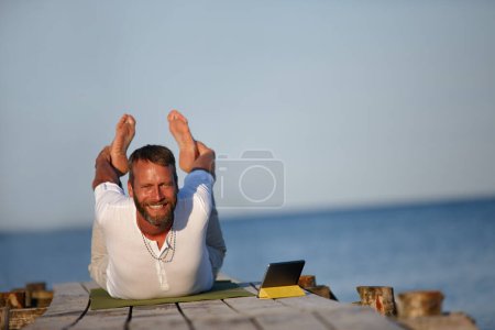 Photo for Mastering the bow position. A handsome mature man doing yoga exercises on a pier with the help of his digital tablet - Royalty Free Image