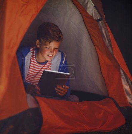 Téléchargez les photos : Accéder à la toile depuis sa tente. un jeune garçon utilisant une tablette numérique en camping - en image libre de droit