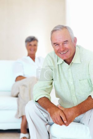 Téléchargez les photos : Homme et femme mûrs souriants dans le salon. Portrait d'homme mûr heureux souriant dans le salon avec une femme en arrière-plan - en image libre de droit