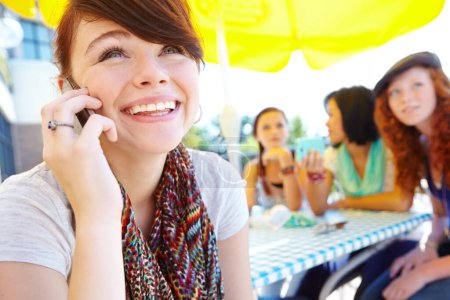 Foto de Es genial saber de ti. Una adolescente sonriendo mientras habla en su celular con sus amigos en el fondo - Imagen libre de derechos