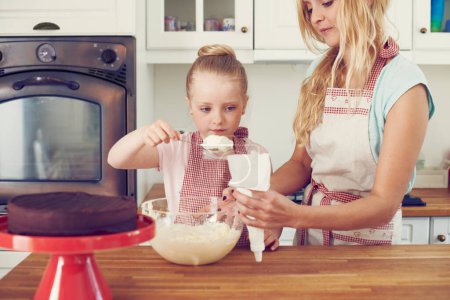 Photo for Getting ready to ice the cake. Cute little girl icing a cake with her mom in the kitchen - Royalty Free Image