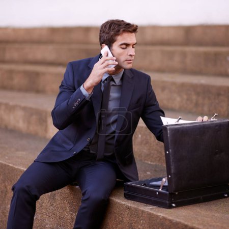 Foto de Multitarea como un profesional en la ciudad. Un hombre de negocios hablando por teléfono mientras está sentado con su maletín en algunos pasos - Imagen libre de derechos