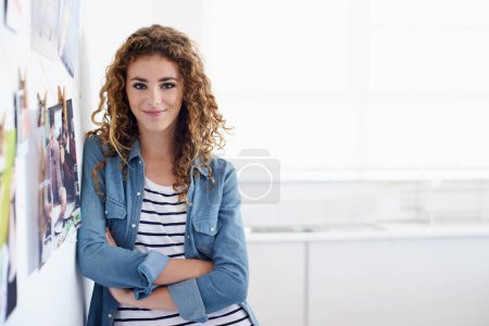Foto de Siguiendo una carrera en diseño gráfico. Una joven diseñadora gráfica sonriendo a la cámara mientras se para junto a un arreglo de fotos en una pared - Imagen libre de derechos