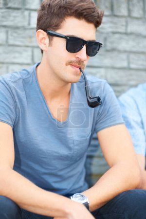 Foto de Haciendo todo con un estilo individual. Guapo joven hipster fumar una pipa mientras está sentado al aire libre - Imagen libre de derechos