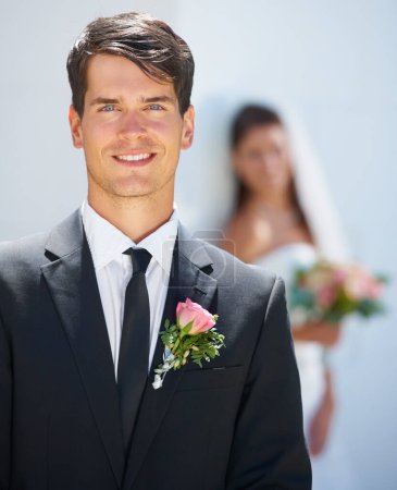 Photo for Her Prince Charming. A handsome young groom smiling at you while his wife-to-be stands blurred in the background - Royalty Free Image