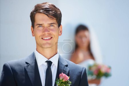Photo for Feeling amazing on this day. A handsome young groom smiling at you while his wife-to-be stands blurred in the background - Royalty Free Image