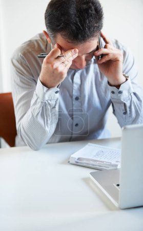 Foto de Estos números no pueden ser correctos... un hombre de negocios agotado hablando por teléfono mientras está sentado frente a su computadora portátil en su escritorio - Imagen libre de derechos
