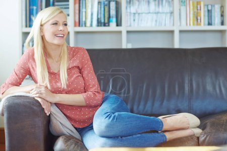 Téléchargez les photos : Belle jeune femme blonde rêvant et se relaxant à la maison sur le canapé. Plein corps d'une femme joyeuse assise sur le canapé dans le salon. Femme confortable souriant tout en se reposant seul. - en image libre de droit