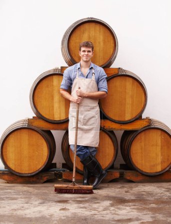Photo for Portrait, broom and oak barrels with a man cleaning the wine cellar of a beverage distillery. Confident, apron and a male cleaner in a winery for the production, fermentation or storage of alcohol. - Royalty Free Image