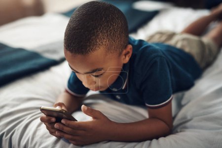 Foto de Aprender es mucho más divertido a través de juegos educativos. un niño usando un celular mientras está acostado en su cama - Imagen libre de derechos