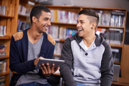 Foto de Riendo y aprendiendo. dos jóvenes riendo mientras usan una tableta digital en la biblioteca universitaria - Imagen libre de derechos