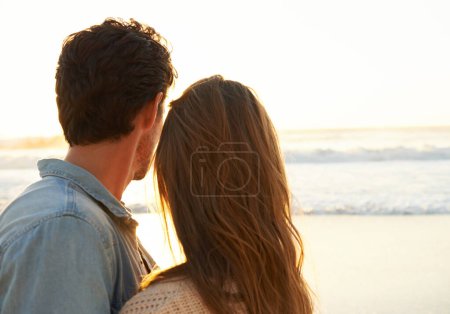 Téléchargez les photos : Une vraie romance. Un jeune couple s'embrassant et regardant le soleil se coucher sur la plage - en image libre de droit
