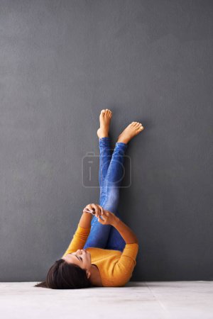 Foto de Mensajería relajada... Una joven usando un teléfono móvil mientras está acostada en el suelo con los pies contra la pared - Imagen libre de derechos