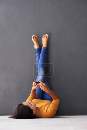 Foto de El enfoque al revés de los mensajes de texto. Una joven usando un teléfono móvil mientras está acostada en el suelo con los pies contra la pared - Imagen libre de derechos