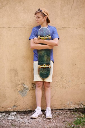 Foto de La fase de monopatín... un adolescente sosteniendo su monopatín mientras está de pie contra una pared - Imagen libre de derechos