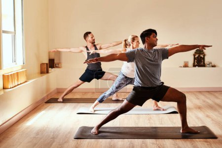 Foto de Dale a tu cuerpo el don del equilibrio. un grupo de jóvenes hombres y mujeres practicando la pose guerrera durante una sesión de yoga - Imagen libre de derechos