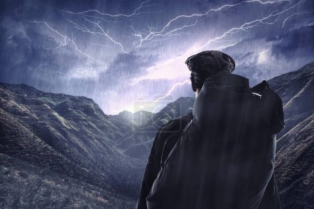 Foto de Tormenta relámpago, viajes y un hombre caminando por la espalda en las montañas durante la noche con lluvia de invierno. Cielo oscuro, nubes grises y clima tormentoso con un caminante masculino al aire libre en condiciones peligrosas. - Imagen libre de derechos