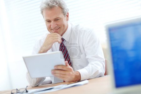 Photo for Receiving great news via email. Mature office employee holding a tablet and smiling while sitting at a desk - Royalty Free Image
