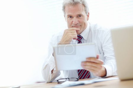 Foto de El negocio digital marca la diferencia. Cabeza y hombros disparados de un hombre de negocios maduro sonriendo ligeramente mientras sostiene una tableta en la oficina - Imagen libre de derechos