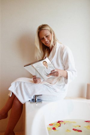 Téléchargez les photos : Mon spa personnel. Une belle jeune femme lisant un magazine alors qu'elle est assise sur le bord d'un bain luxueux rempli de pétales - en image libre de droit