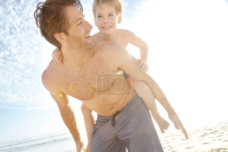 Téléchargez les photos : Obtenir un piggyback de papa. un jeune père donnant à son fils une balade à dos de cochon à la plage - en image libre de droit