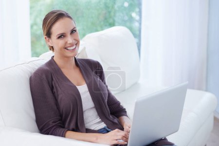 Téléchargez les photos : Connectivité instantanée. Une jeune femme souriante assise sur son canapé avec son ordinateur portable - en image libre de droit