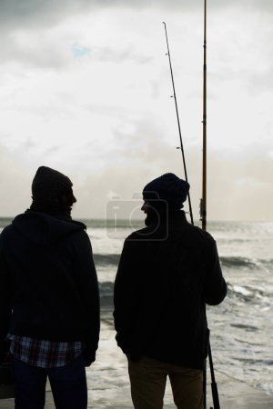 Téléchargez les photos : S'amuser lors d'un voyage de pêche. deux jeunes hommes pêchant à l'océan tôt le matin - en image libre de droit