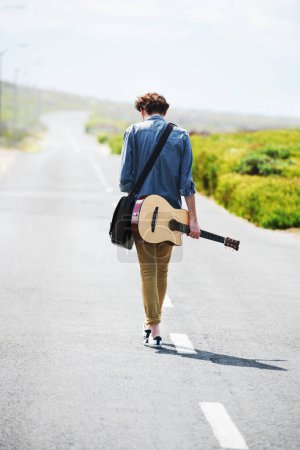Foto de Llevando mi música a la carretera. Vista trasera de un joven hombre de moda caminando por el camino sosteniendo una guitarra acústica - Imagen libre de derechos
