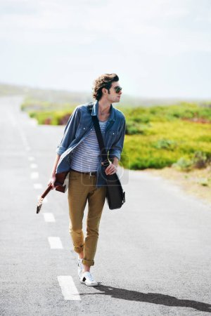 Photo for Getting some musical inspiration on the open road. Trendy young male wearing sunglasses walking down the middle of a road holding a guitar - Royalty Free Image