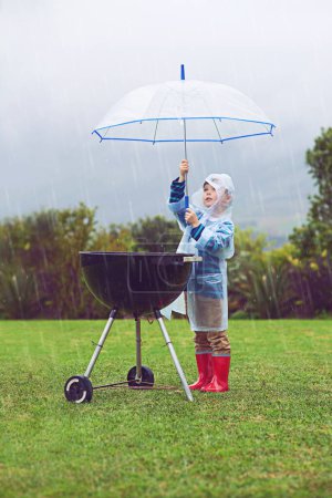 Foto de Lluvia, paraguas y un niño cubriendo una barbacoa al aire libre en un campo mientras cocina comida sola en invierno. Impermeable, niños y seguro con un niño pequeño tratando de proteger una barbacoa durante el clima tormentoso. - Imagen libre de derechos