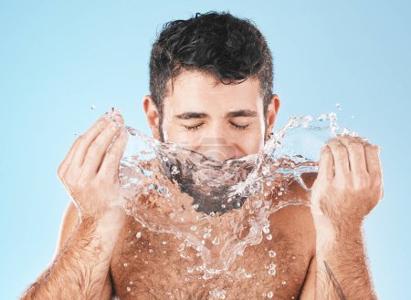 Téléchargez les photos : Man, splash and face for beauty in studio for wellness, health or self care cleaning by blue background. Model, bathroom and water drops with hands for healthy facial, self love and cosmetic backdrop. - en image libre de droit