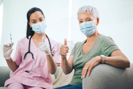 Photo for More than a doctor...Portrait of a masked doctor and her masked patient showing the thumbs up after receiving an injection at home - Royalty Free Image