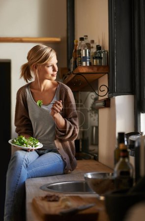 Téléchargez les photos : Moments confortables dans la cuisine. Plan complet d'une jeune femme mangeant un bol rempli de légumes feuillus dans une cuisine rustique - en image libre de droit