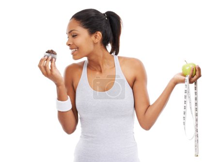 Photo for Its just so tempting...A pretty young woman choosing a cupcake over an apple and a measuring tape - Royalty Free Image